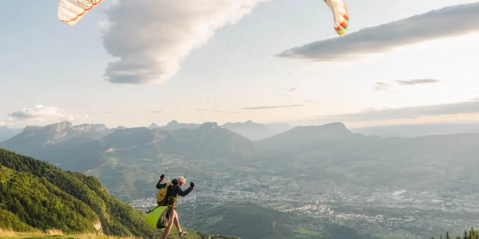 Un parapentiste prend son envol en parapente sur l’aire de décollage du Sire en Savoie, avec les reliefs et la vallée en arrière-plan.